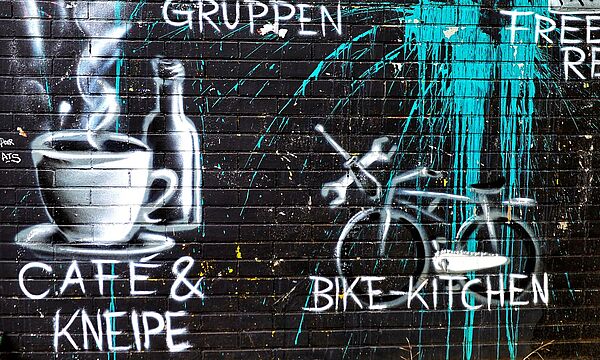 Graffitti auf einer Wand: u.a. Kaffeetasse, Farrad und Worte: z.B. Café und Kneipe, bike-kitchen, Gruppen